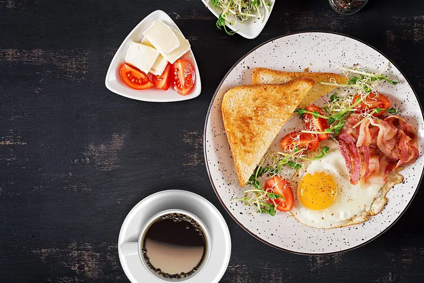 A breakfast plate of toast, bacon, tomato and a fried egg with a cup of coffee